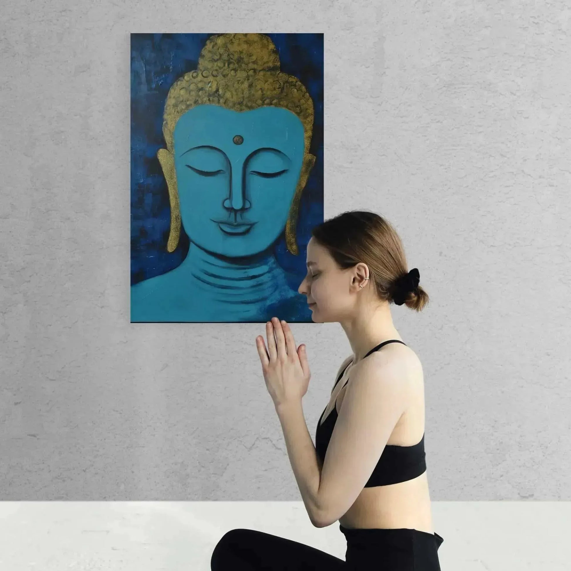 A woman in a yoga pose kneels with her hands in prayer, radiating Zen calmness while facing a gray wall. Above her, the Tranquil Buddha Head Canvas meditation yoga pose painting from Buddha in Art Oregon by Printify adds serenity to the scene. She is dressed in a black sports bra and leggings, with her hair neatly tied back.