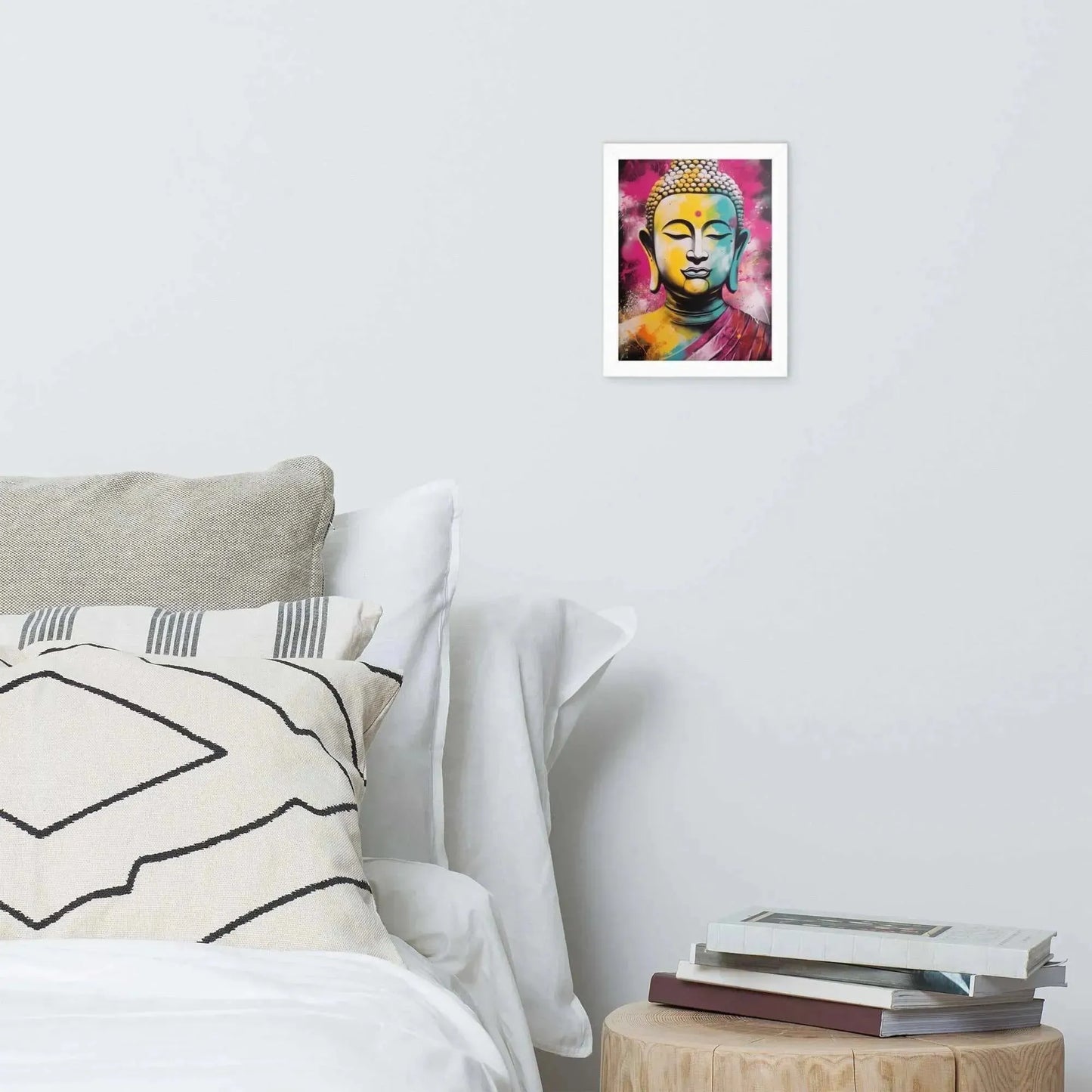 A vibrant framed poster of a stylized Modern Buddha Painting hangs on a white wall above a neatly arranged bed with plush pillows and a decorative cushion. A bedside table to the right holds a stack of books beside a white bed covered in crisp linen. A wooden tray with a black bowl and a jar rests on the bed, adding a homely touch to the modern, serene bedroom setting.