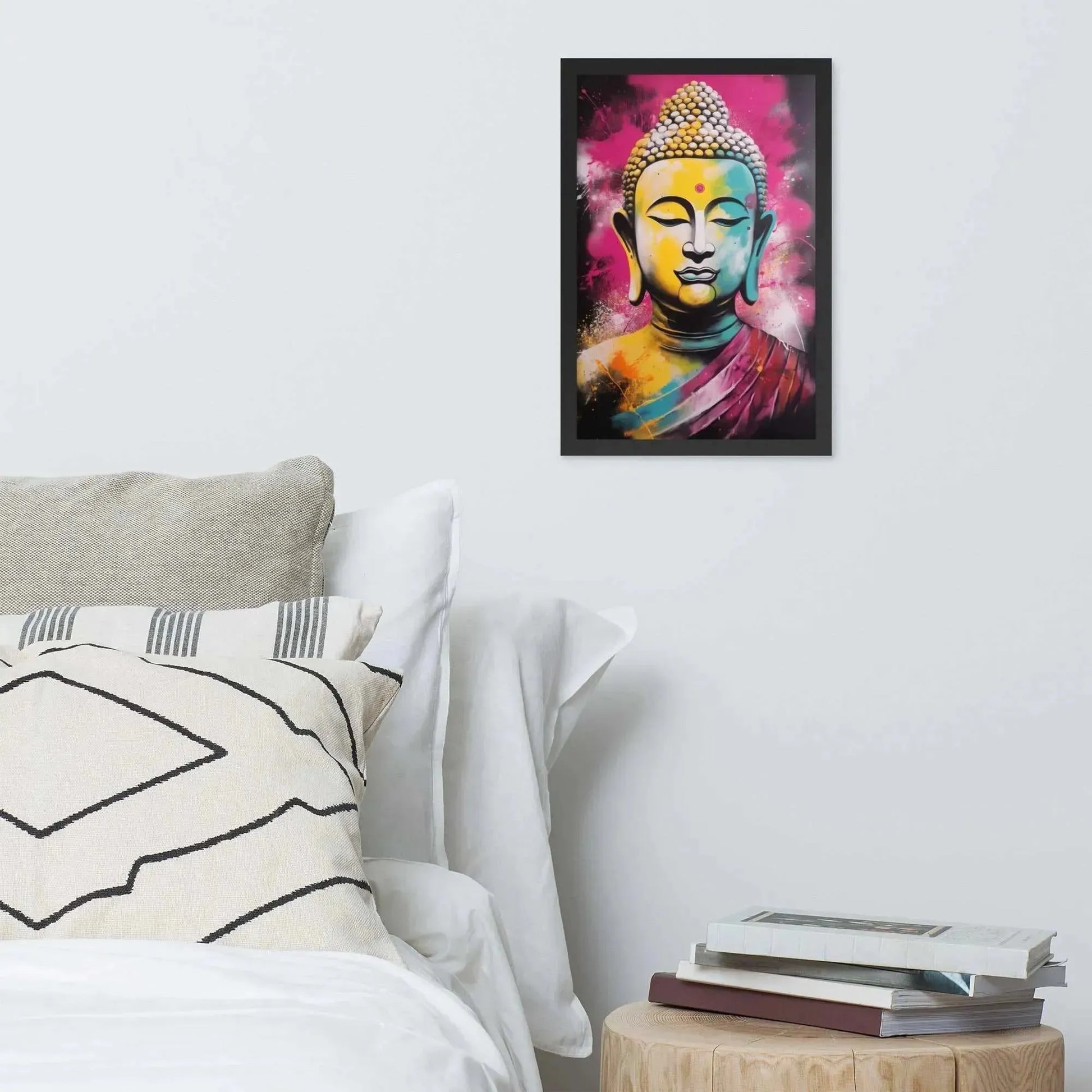 A vibrant framed poster of a stylized Modern Buddha Painting hangs on a white wall above a neatly arranged bed with plush pillows and a decorative cushion. A bedside table to the right holds a stack of books beside a white bed covered in crisp linen. A wooden tray with a black bowl and a jar rests on the bed, adding a homely touch to the modern, serene bedroom setting.