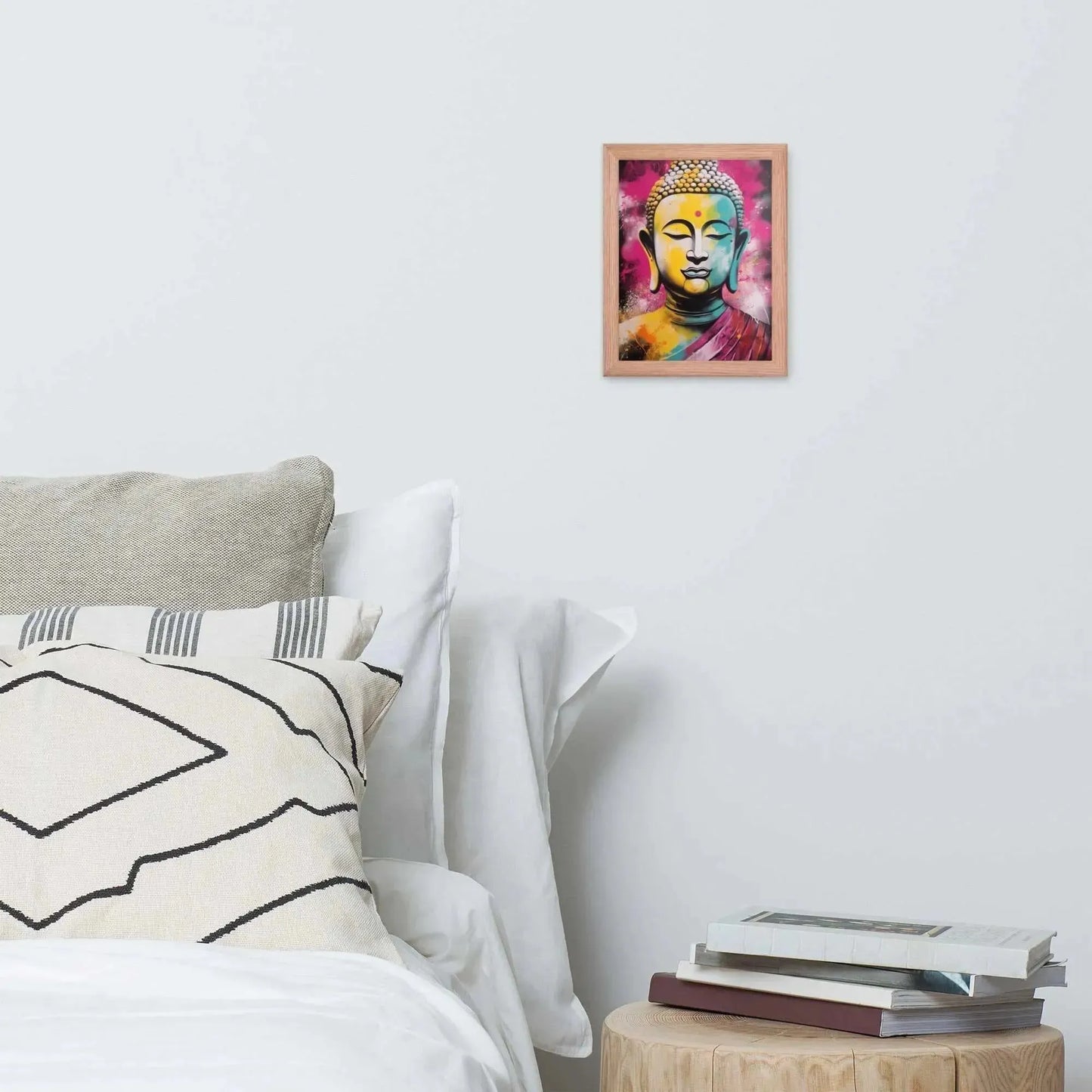 A vibrant framed poster of a stylized Modern Buddha Painting hangs on a white wall above a neatly arranged bed with plush pillows and a decorative cushion. A bedside table to the right holds a stack of books beside a white bed covered in crisp linen. A wooden tray with a black bowl and a jar rests on the bed, adding a homely touch to the modern, serene bedroom setting.