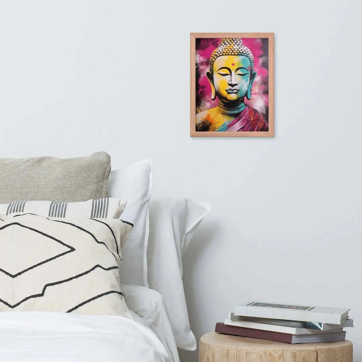 A vibrant framed poster of a stylized Modern Buddha Painting hangs on a white wall above a neatly arranged bed with plush pillows and a decorative cushion. A bedside table to the right holds a stack of books beside a white bed covered in crisp linen. A wooden tray with a black bowl and a jar rests on the bed, adding a homely touch to the modern, serene bedroom setting.