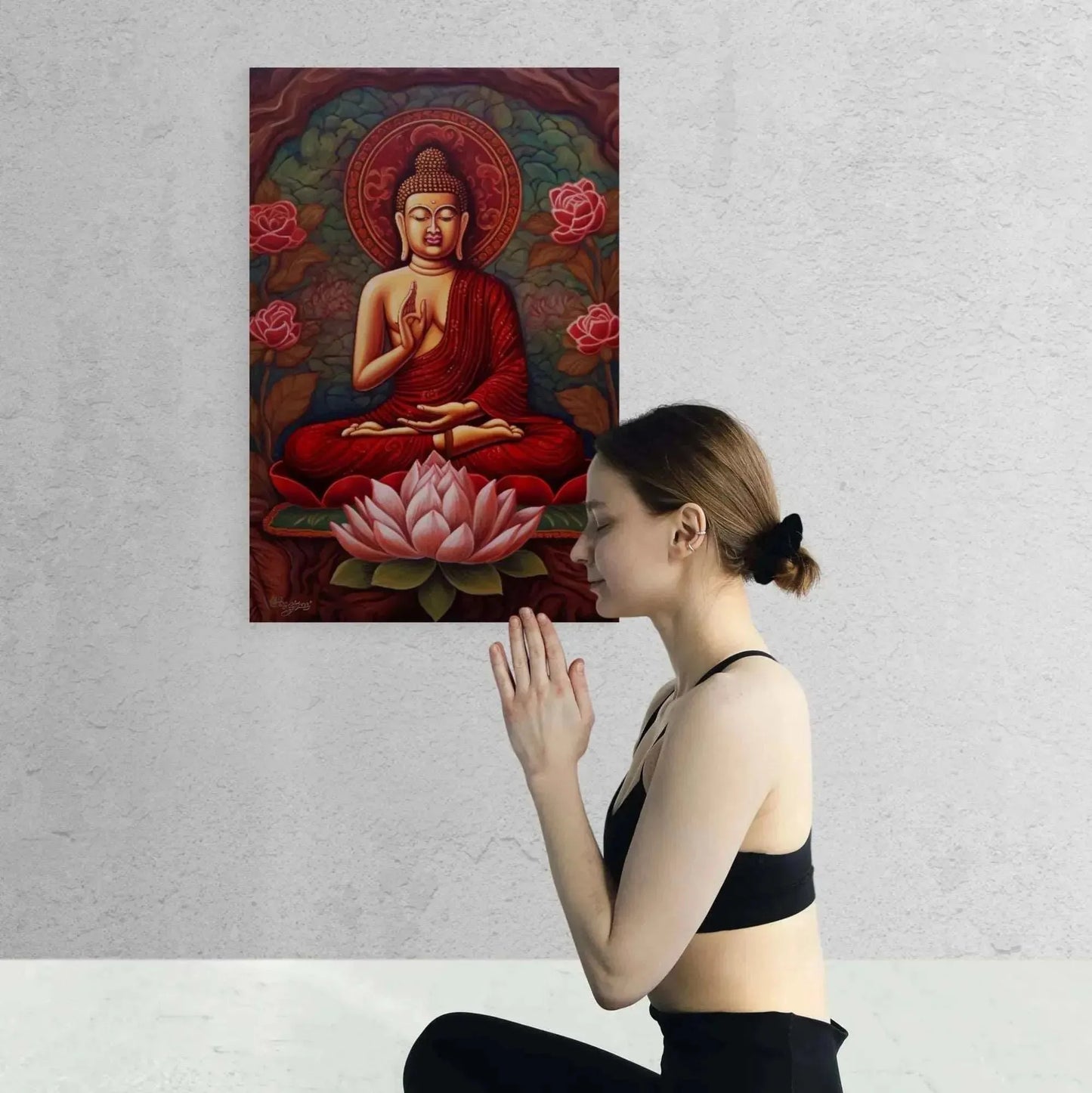 A woman in athletic wear kneels in meditation with folded hands before the "Sitting Buddha Print: A Striking Abstract from ZenArtBliss.com" by Printify, depicting a Buddha sitting on a lotus surrounded by lotus flowers. The background is a plain, light-colored wall, enhancing the calming essence of this Buddhist-themed art.